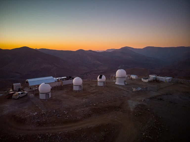 Fotografía panorámica del observatorio El Sauce: Se observan 4 cúpulas blancas y otros edificios pequeños que contienen los telescopios, en un paisaje desértico al atardecer.